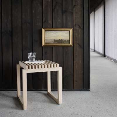 Perfekt für die Wasserkaraffe: Skagerak Cutter Hocker aus hellem Holz vor einer dunklen Wand. 